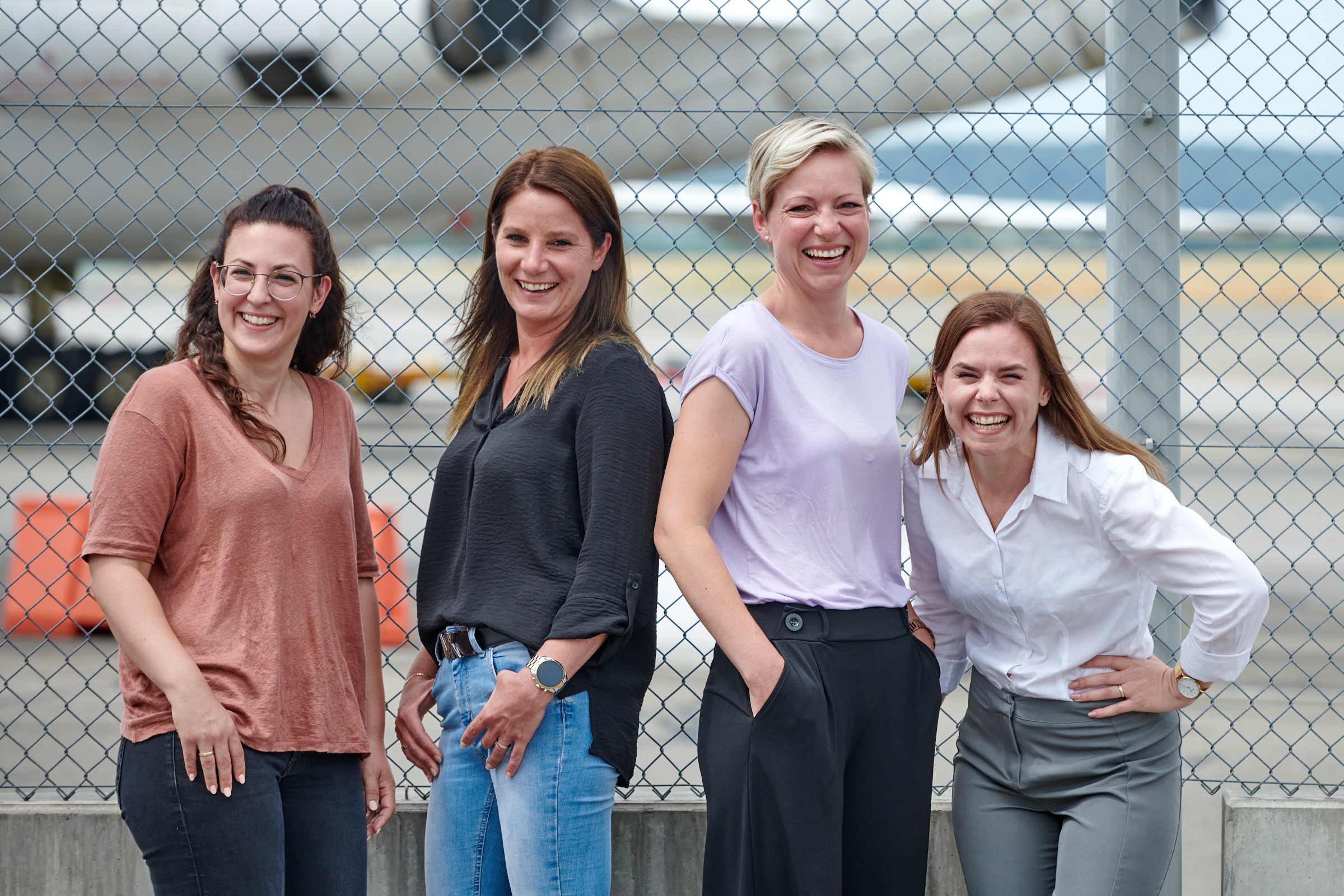 gruppenbild-mit-vier-frauen-lachend-vor-zaun-am-flughafen
