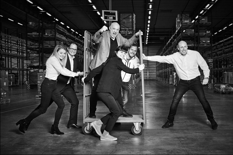 gruppenbild-team-schiebt-wagen-in-logistikhalle-chef-schreit
